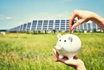 A hand putting money into a piggy bank with solar panels in the background, indicating solar panels are very expensive.